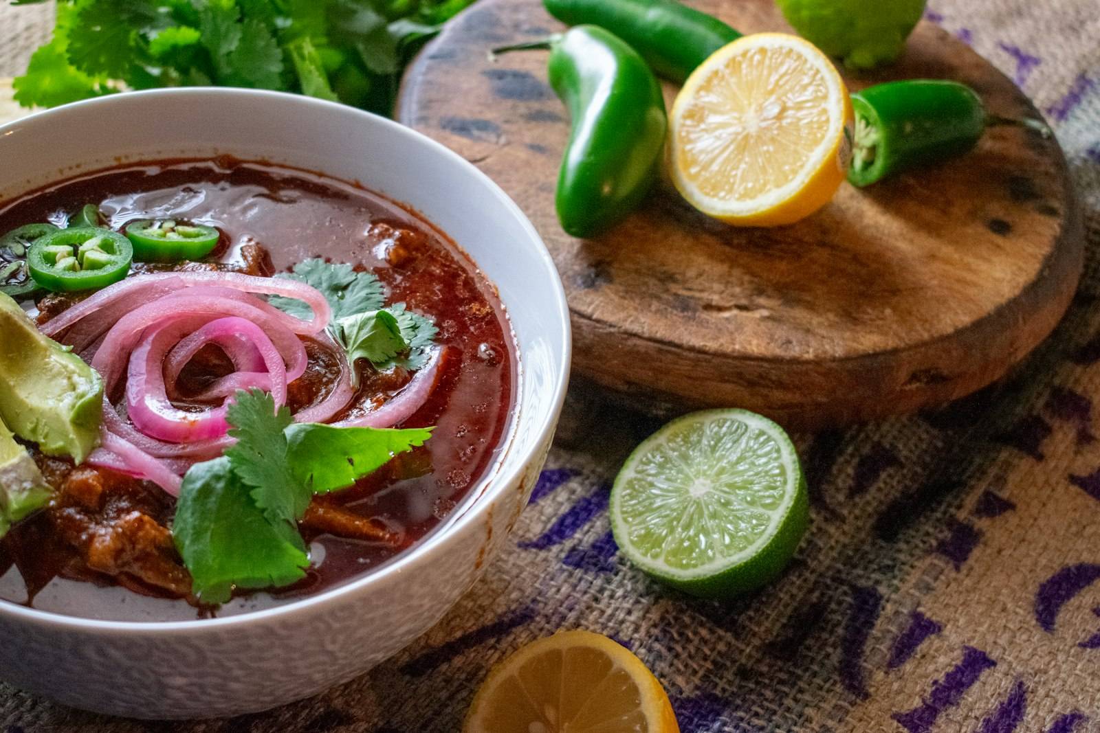 A bowl of red consomé with pickled onion, cilantro, jalapeño and avocado