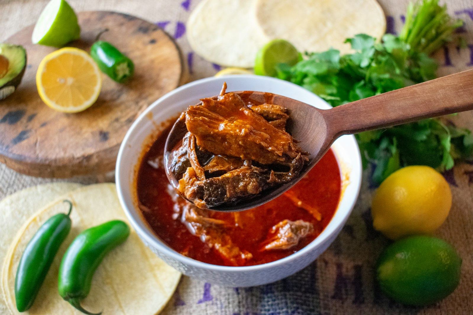 A ladle lifting birria out of a bowl of red broth