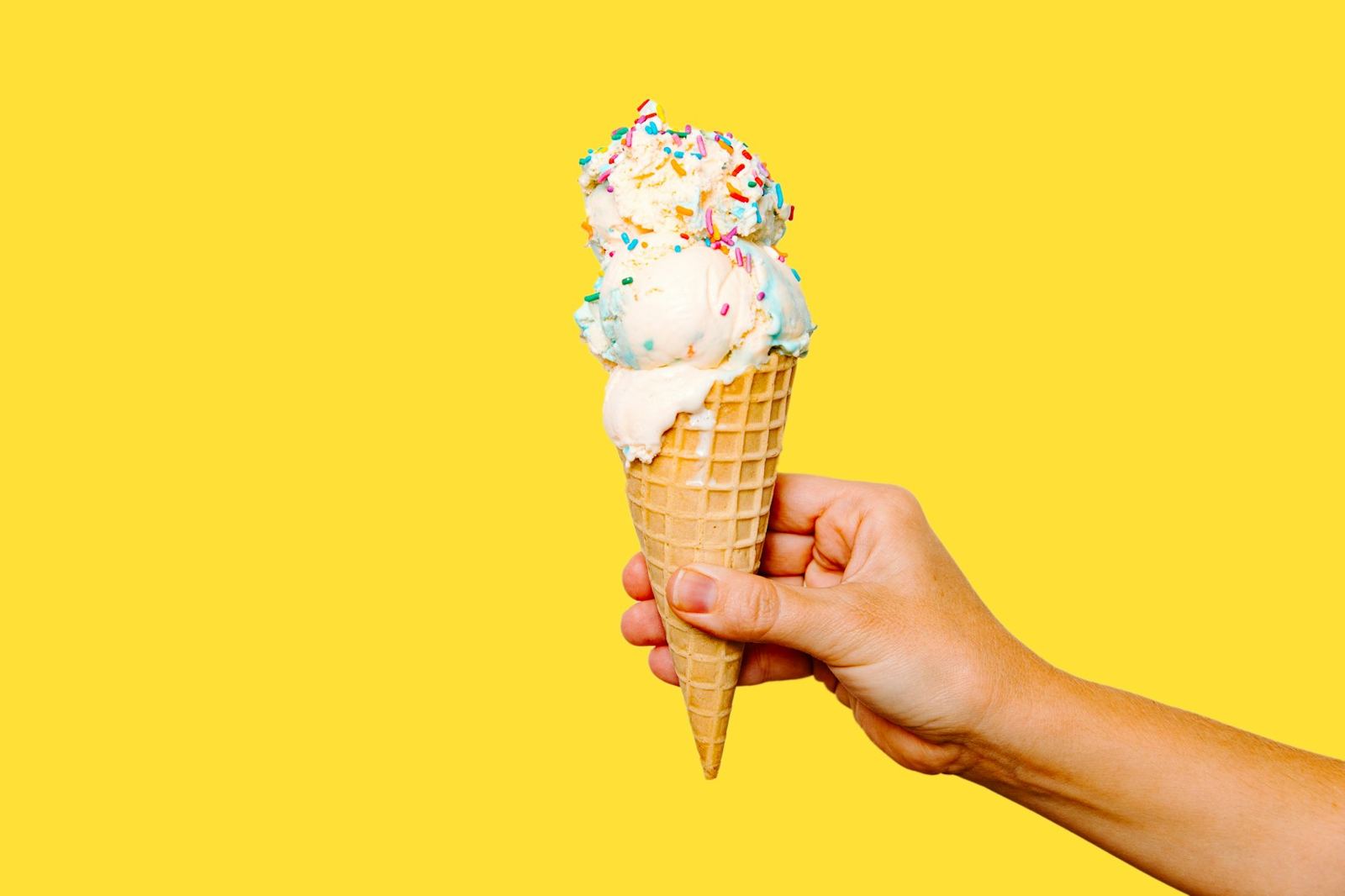 A hand holding a sprinkle-topped cone against a yellow wall
