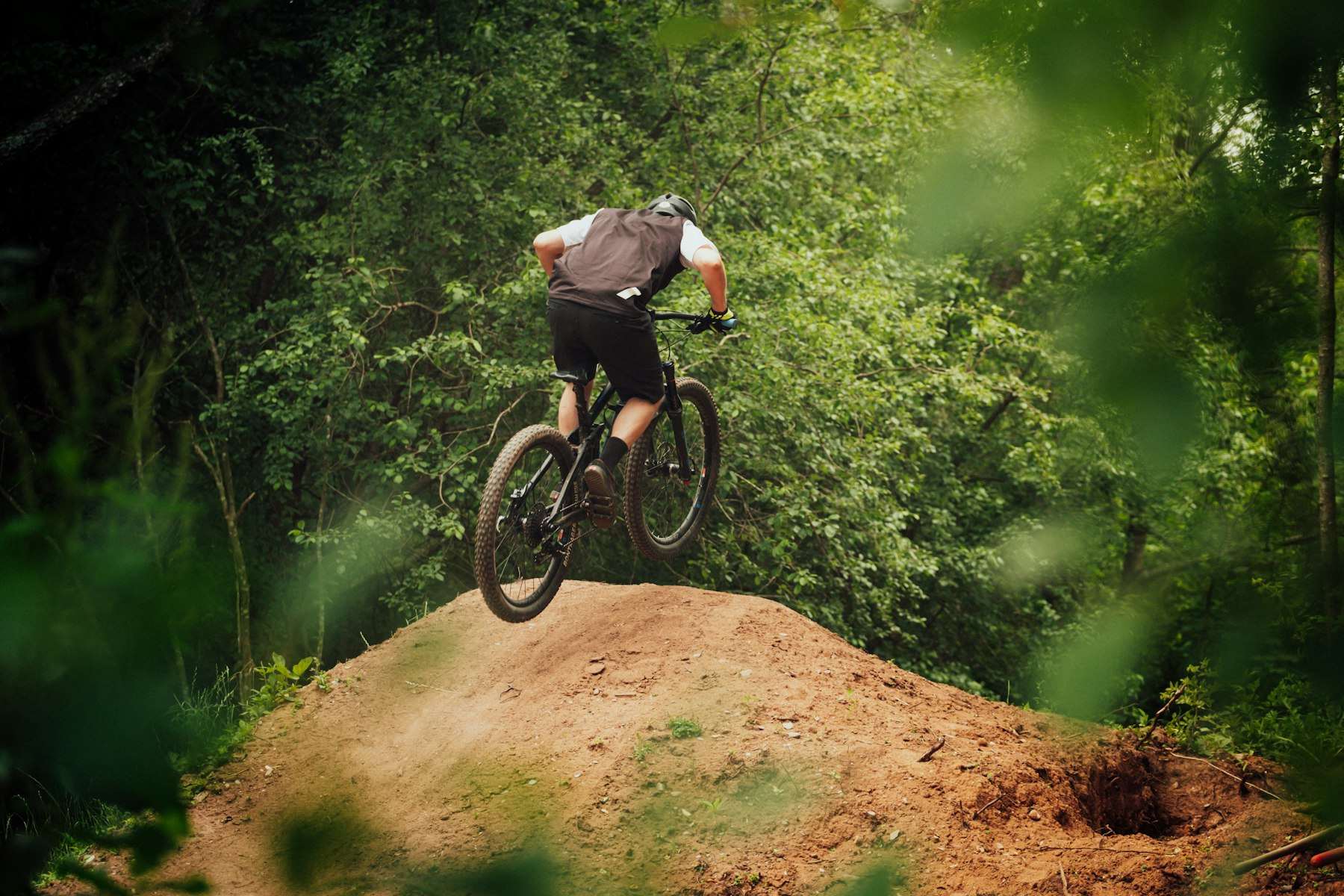 Man in black jacket riding black mountain bike on brown dirt road between green trees during